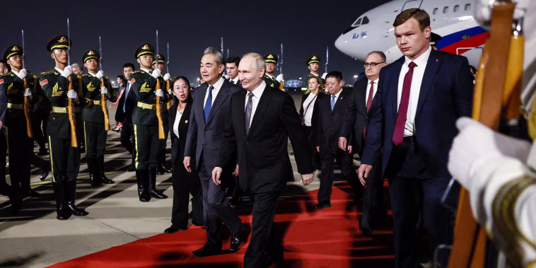 Vladimir Putin and Wang Yi walk along a red carpet extending from a plane parked in the background, with soldiers and other officials surrounding them