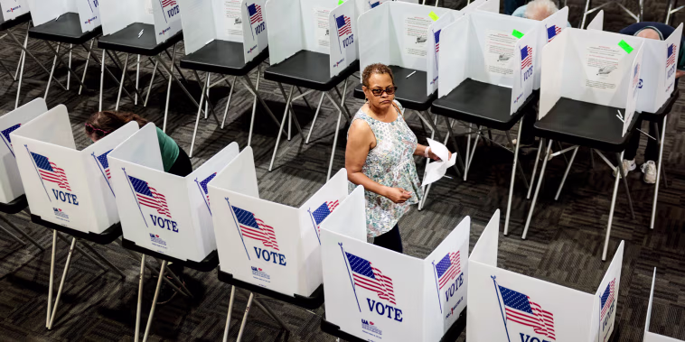 Pamela Dear walks down an aisle between rows of voting booths inside of a room