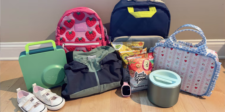 Back-to-school gear for kids laid out on a wood floor: two backpacks (pink strawberry print and navy), a green lunchbox, white toddler sneakers, a folded jacket, Little Spoon snack pouches, a kids' smartwatch, an insulated food container, and a floral gingham lunch bag.