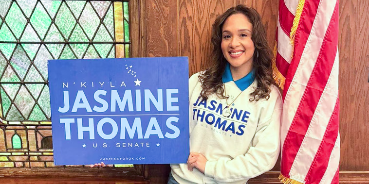 N’Kiyla Jasmine Thomas stands in front of a campaign table while holding a blue sign that reads "N’Kiyla Jasmine Thomas, U.S. Senate"