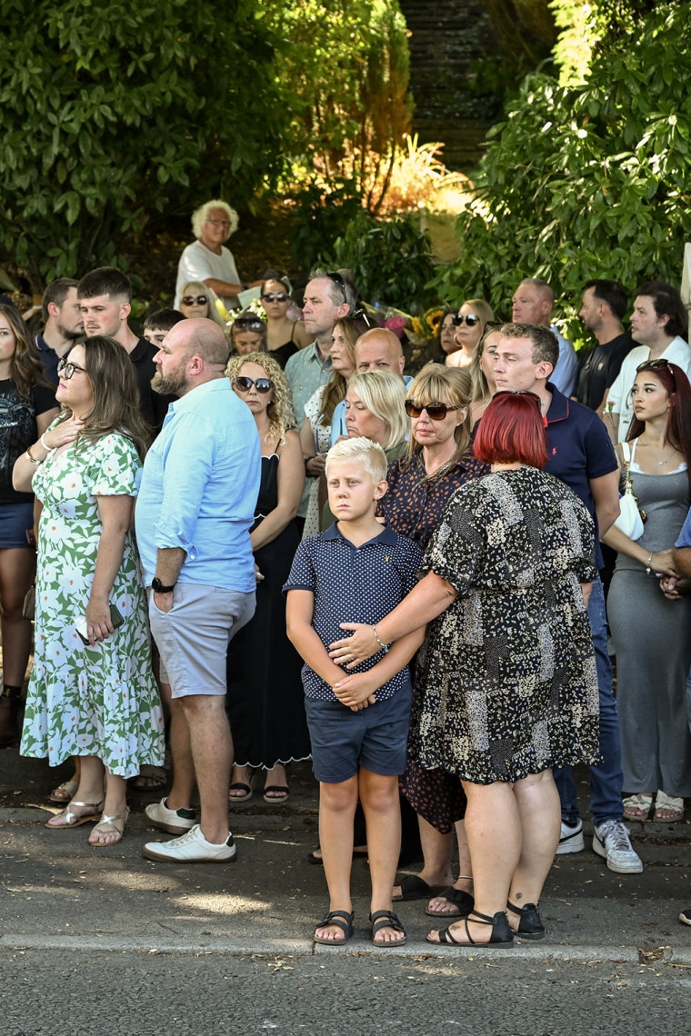 Dozens of people standing along a shaded street.