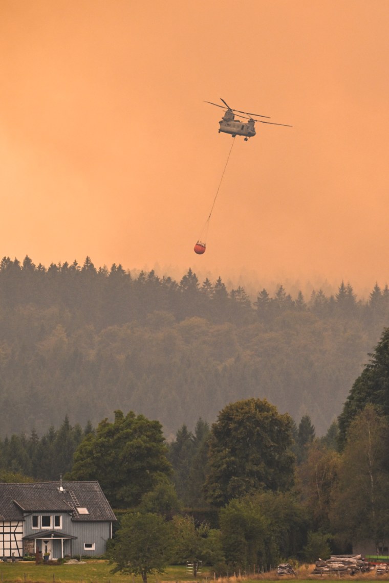 A Royal Netherlands Air Force Boeing CH-47D Chinook carrying a bambi bucket through an orange smokey sky over a forested village