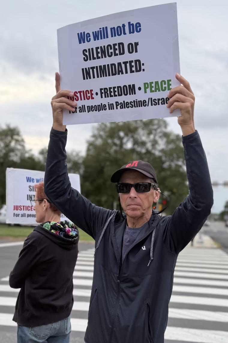 A man in sunglasses holds a sign reading "we will not be silenced or intimidated".