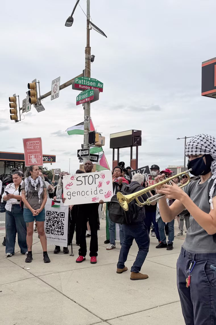 A crowd of protestors, one with a sign reading STOP GENOCIDE, gathered in front of a metro station.