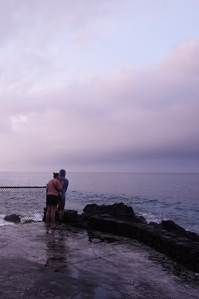 A couple observing a purple sunset over a cloudy sky on the Hawaiian coastline.