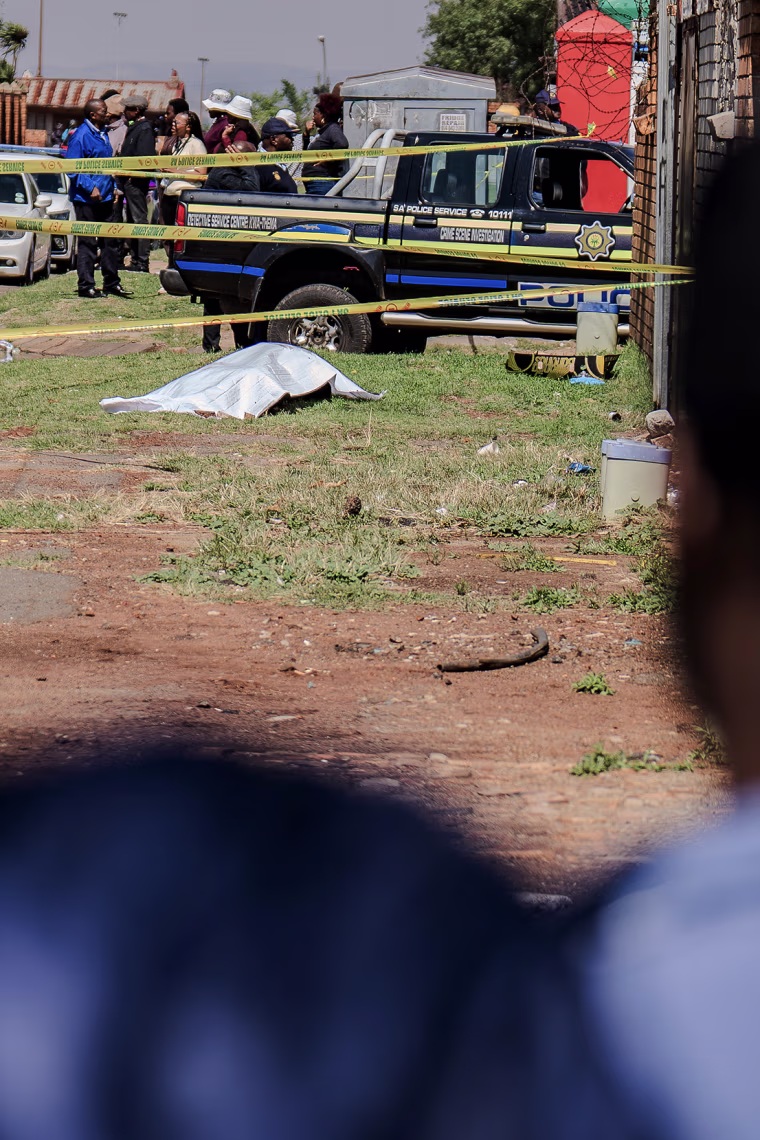 Crime scene tape surrounds a body covered by a gray tarp as people look on from a distance