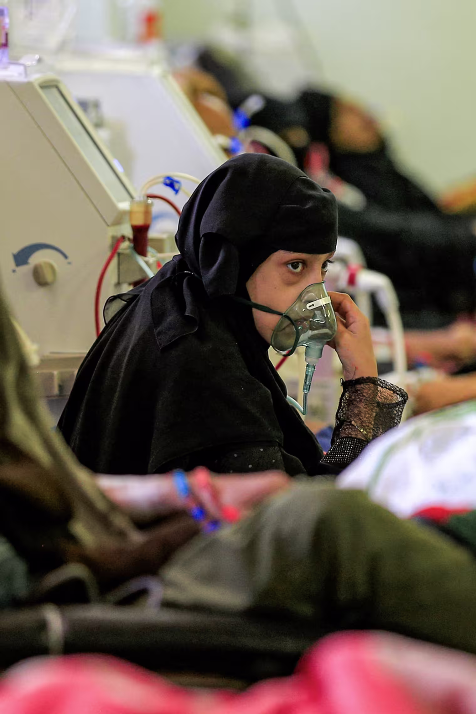 A young girl holds a respirator mask over her face inside a crowded hospital room.