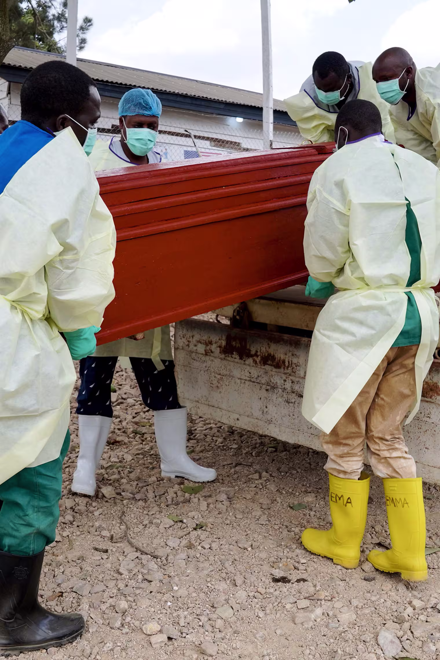 Healthcare workers in disposable scrubs hoist a red coffin into the back of a pickup truck.