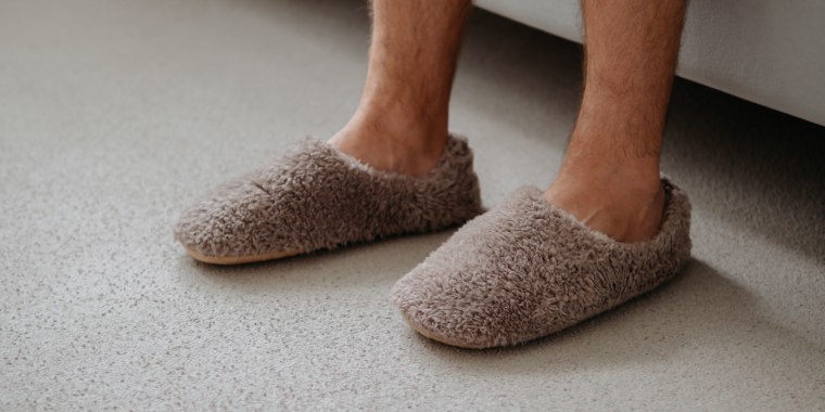 Cropped shot of unrecognizable man in soft plush warm slippers, sitting on sofa in morning time at cozy scandinavian home. Hairy legs of adult man in trendy warm slippers on floor