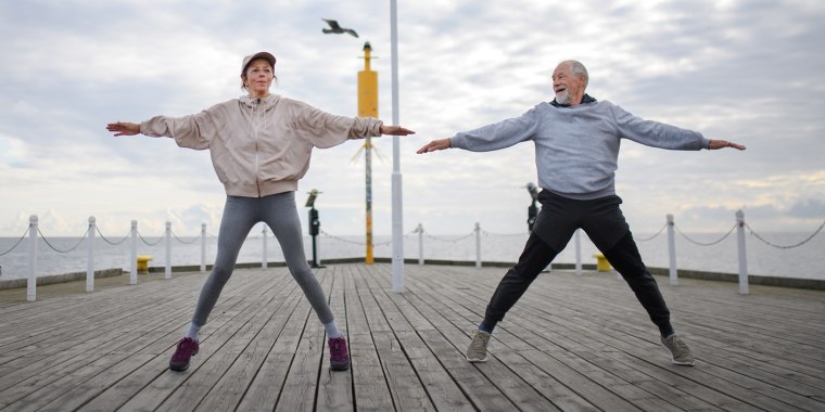 Active senior couple doing jumping jack exercise outdoors on pier by sea in early morning.