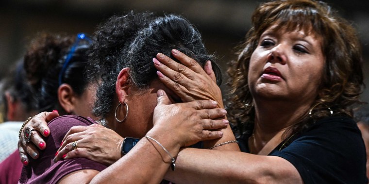 A woman in a maroon shirt covers her face as she cries as another woman in black hugs her