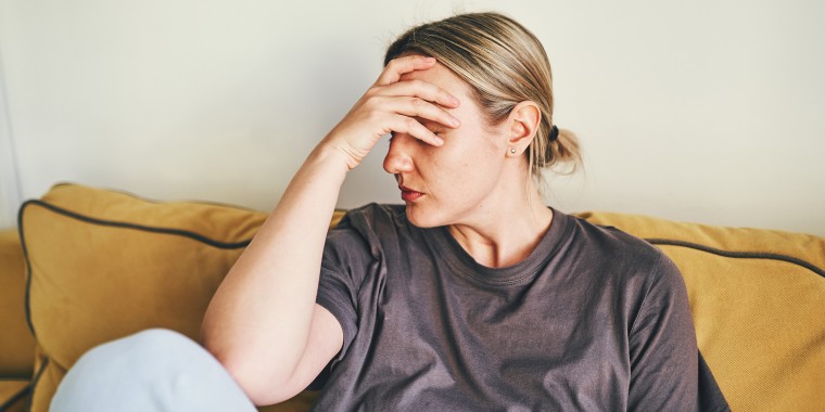 A young woman is sitting at home on a yellow sofa with her head in her hands, showing a headache.