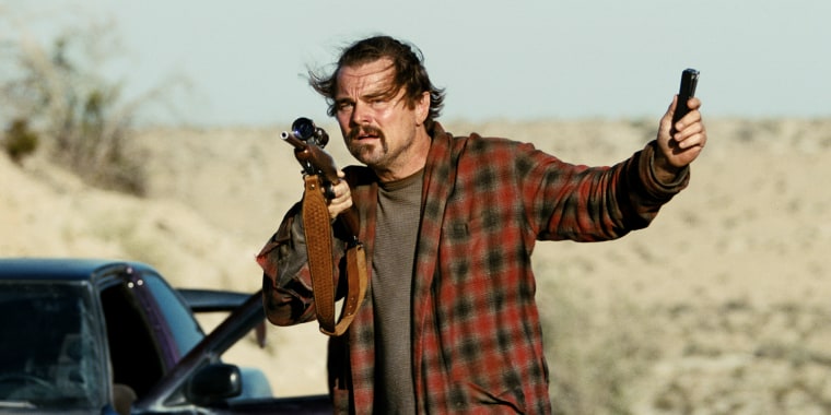 Leonardo Di Caprio stands in a robe on the street holding a gun.