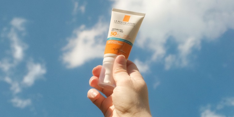 A hand holds tone bottle of sunscreen against a blue sky with clouds