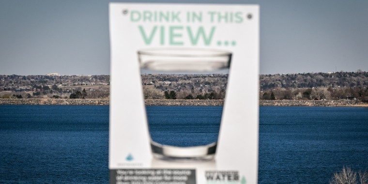A sign with a water glass on it appears in soft focus in front of a large mountain lake. 