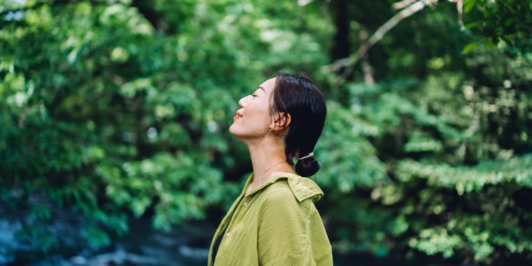 Beautiful young Asian woman meditating in the nature with her eyes closed, setting herself free and feeling relieved. Enjoying fresh air and breathing in the calmness with head up. Freedom in nature. Connecting with nature. Health and wellness concept