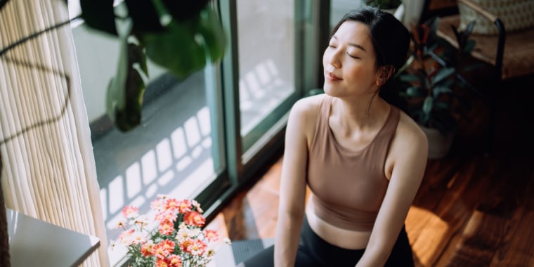 Active young Asian sports woman taking a break after working out at home, sitting on exercise mat taking a deep breath with her eyes closed. Sports and exercise routine. Health, fitness and wellness concept