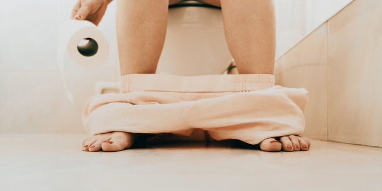 Woman sitting on toilet with pants down holding toilet paper in a bright bathroom
