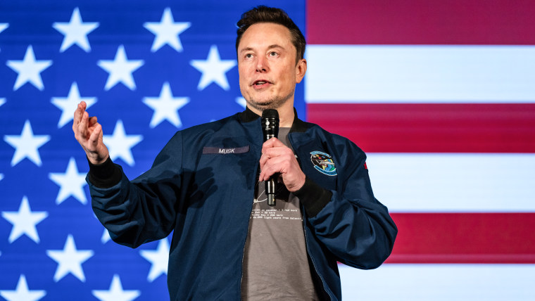 Elon Musk speaks on stage, with an American flag backdrop behind him