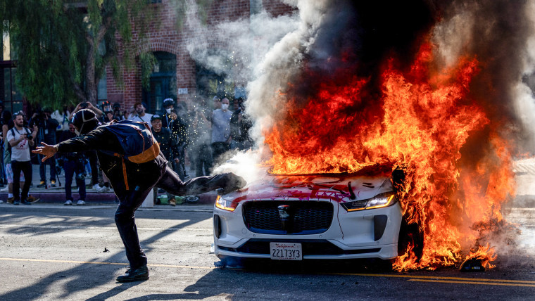 A protester kicks a burning Waymo vehicle outside