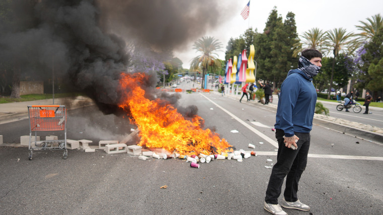 A fire burns as a protester stands across the way from Border Patrol personnel in riot gear and gas masks outside an industrial park in the city of Paramount, south of downtown Los Angeles, on Saturday, June 7, 2025. 
