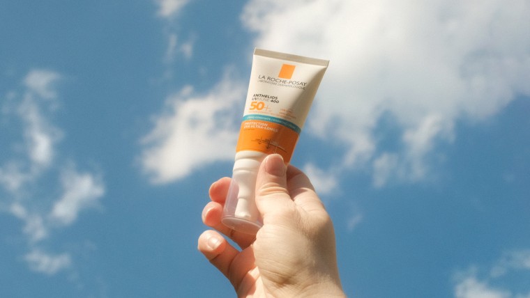 A hand holds tone bottle of sunscreen against a blue sky with clouds