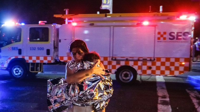 A woman walks outside carrying a child wearing an emergency blanket, an ambulance is on the street behind them