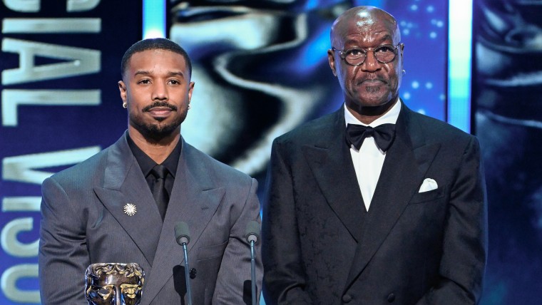 Michael B. Jordan and Delroy Lindo stand at a lectern on stage during an award ceremony. They look nonplussed.