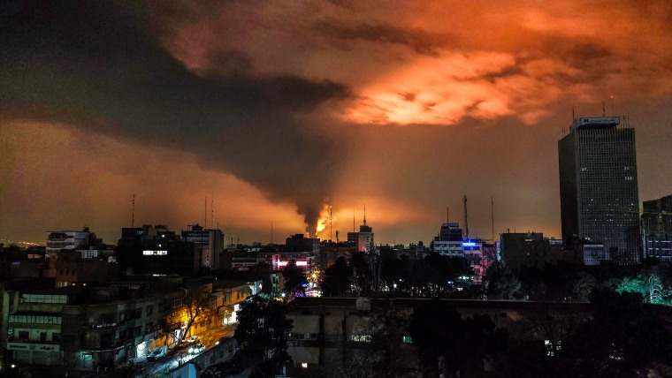 Cloudy, smoky night skies are lit with orange light and flames from distant airstrikes, seen across the rooftops of Tehran.