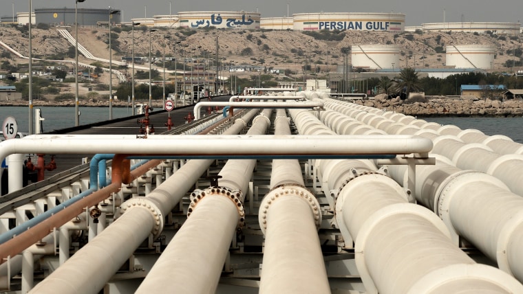 White pipelines in a row lead toward the shore where oil storage tanks are visible in the distance.