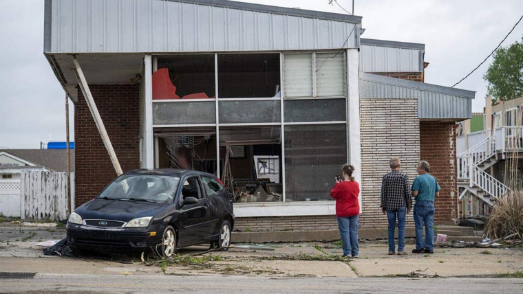 People stand outside of a damaged building with a damaged car parked out front