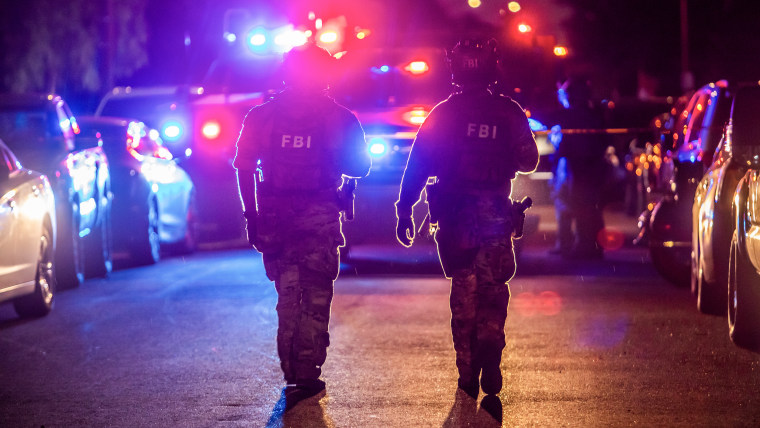 Two FBI field agents wearing helmets and camouflage fatigues walk in front of a vehicle with red and blue flashing lights.