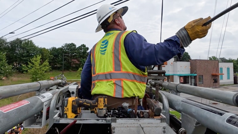 An AT&T technician works on a telephone pole, wearing a light green hazard vest with the AT&T logo and a white hard hat
