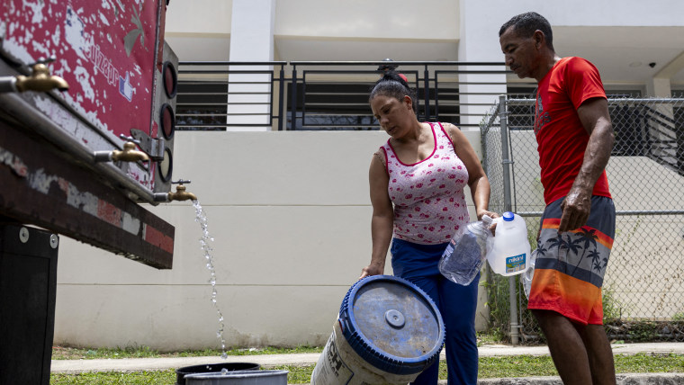 A man and woman collect water from a tanker truck with buckets and plastic bottles.