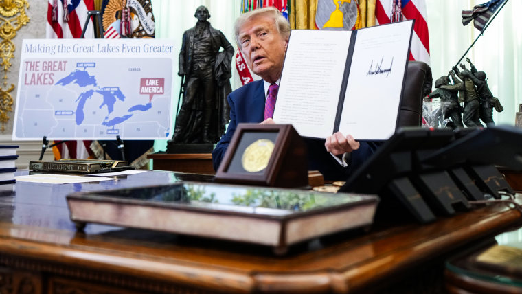 President Donald Trump displays a signed executive order while seated at his desk in the Oval Office, next to him is a poster showing a map of the great lakes that reads "Making the Great Lakes even greater" with Lake Ontario labeled as Lake America