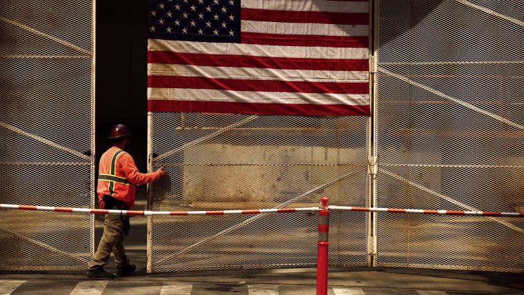 A construction worker wearing an orange vest and hardhat stands beneath a large American flag on a fence.