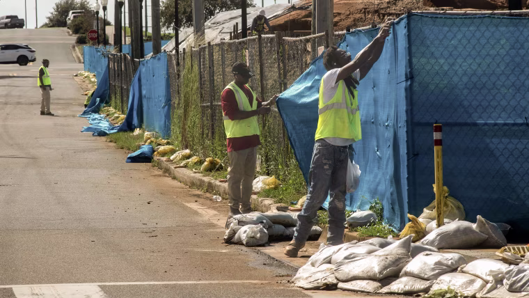 Workers in yellow vests removing blue tarp from a chainlink fence.