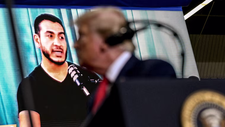 Donald Trump appears in soft focus in front of a digital display of Abdul El-Sayed during a political rally.