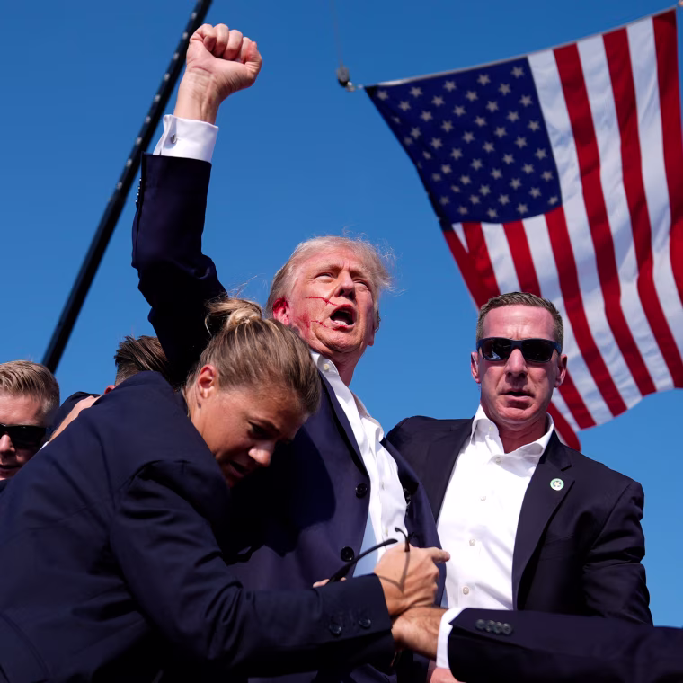 Donald Trump, with blood on his ear, raises up a fist while being escorted away by secret service outdoors, American flag flies in the background