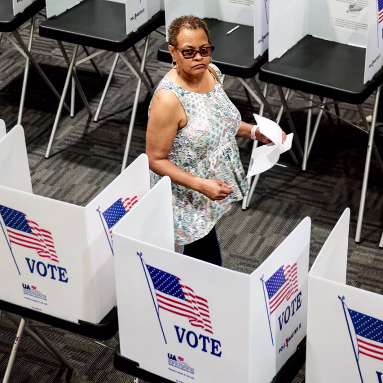 Pamela Dear walks down an aisle between rows of voting booths inside of a room