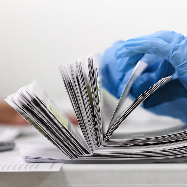 A gloved hand (blue) counts a stack of mail-in ballots on a table, side view
