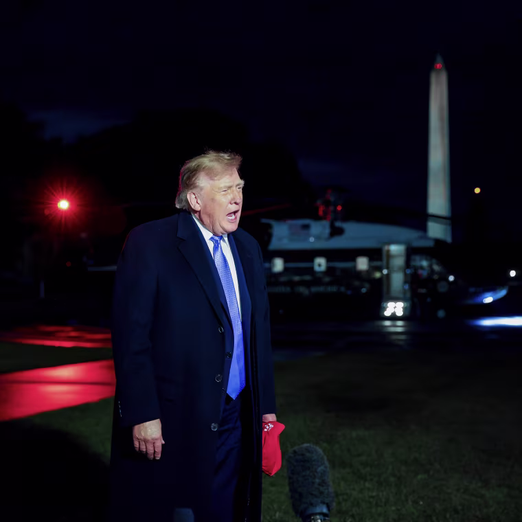 Donald Trump standing in the dark, illuminated by television news camera lighting.
