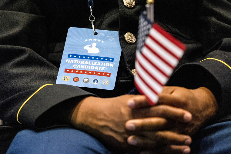 A person seated, focus is on a badge they're wearing that reads "naturalization candidate" as they hold an American flag