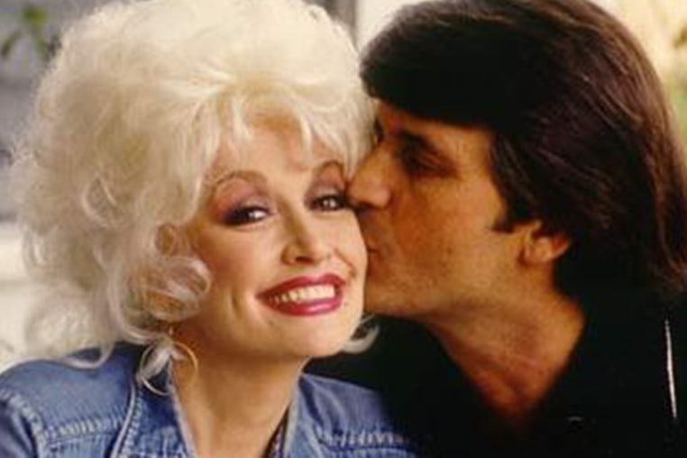 Dolly parton smiles as a dark haired main kisses her cheek.
