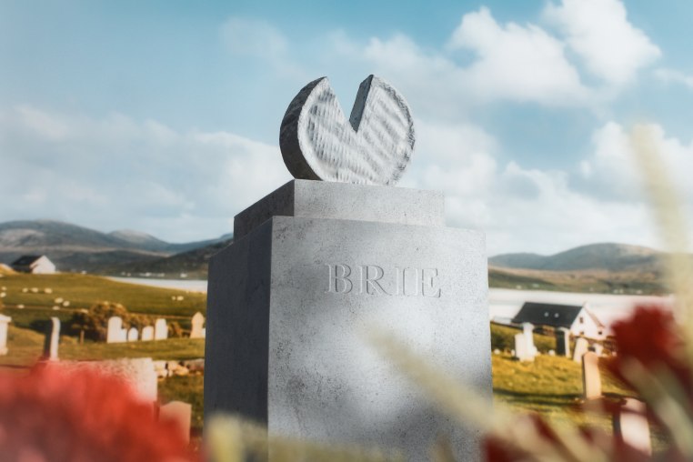 A wheel of cheese shaped gravestone in a cemetery on a hill, engraved with "Brie."