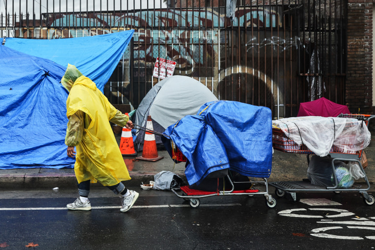 A person walks with carts in the rain near an encampment of unhoused people