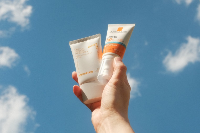 A hand holds two bottles of sunscreen against a blue sky with clouds