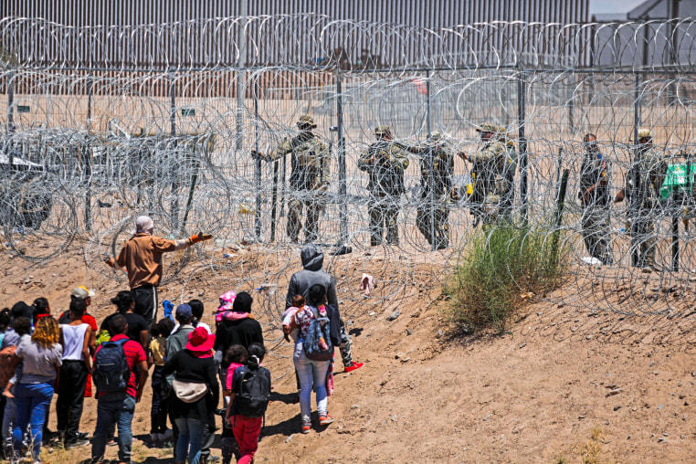 Migrants stand in front of a barbed wire fence opposite from National Guard agents