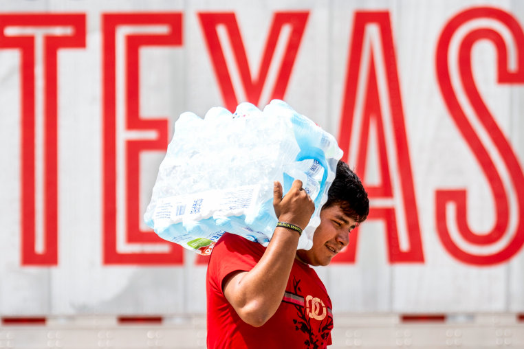 A man carries water on his shoulder in front of a wall with "Texas" written in large red text