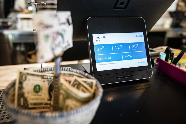 A Square payment device displays tipping options at a coffee shop next to a basket of dollar tips